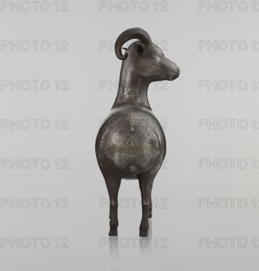 Ram, Persian, Qajar dynasty 1785–1925, early 19th century 1st half of the 19th century, Iran. Sculpture; animal figurine of a ram. Materials/technique: decorated with geometric motifs and floral ornament; back decorated with medallions and palmettes with floral ornament. Dimensions: height 22.5 cm, width 20.5 cm, depth 9.0 cm; weight 0.64 kg. Creator: unknown., A sculptural object resembling an animal with curved horns stands on a smooth reflective surface against a pale gray background, its form elongated with a rounded torso and slender legs; the surface of the object is primarily dark brown to black with areas of lighter brown and subtle gray wear, and it is decorated with several ornate swirling and geometric patterns in muted gold and faded yellow tones, with additional fine line markings on the head and neck in similar light colors.