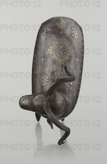 Ram, Islamic art, Persian Qajar dynasty, 1785–1925, 1st half of the 19th century, Iran; sculpture; animal figurine ram with horns; decorated on the back with medallions and palmettes with floral ornament; geometric motifs; materials not specified; height 22.5 cm, width 20.5 cm, depth 9.0 cm, weight 0.64 kg., A sculptural object resembling an elongated animal form sits upright on a reflective surface; it is primarily dark gray with a weathered metallic texture and has decorative motifs in muted gold and lighter gray. The object features a rounded body with a tapered head-like end and curving horn- or leg-like appendages extending from the lower portion, all sharing the same dark gray tone. Several ornate patterns in gold appear on the top surface and along the sides, including symmetrical medallion shapes and small floral or starlike embellishments in light gray. The reflective surface below shows a soft mirror image of the object in pale gray. The background is a smooth, even light gra...