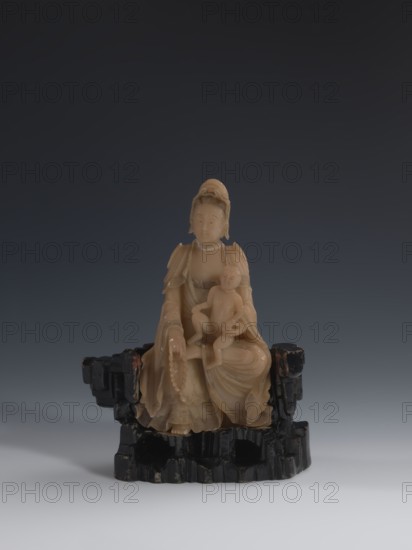 Guanyin Giving Birth to a Son on a Rock, sculptor unknown. Figurine of the goddess made of light coffee-colored alabaster, on a stone base field spar with red and pink spots, painted with black lacquer. Goddess holds a rosary in her right hand and a child in her left. Chinese, Qing dynasty 1644–1911, 18th–19th century. Sculpture figurine. Height 18.2 cm; width 13.8 cm., A carved figurine in a warm beige or light tan material sits on a dark, almost black irregular base; the figurine depicts a seated robed figure holding a smaller childlike figure on a lap, with flowing drapery details and a beaded strand hanging from a hand, both figures showing smooth carved facial features and hair details, while the base is textured and blocky with worn edges and traces of reddish brown on some raised areas, all set against a gradient background ranging from deep charcoal at the top to pale gray at the bottom.