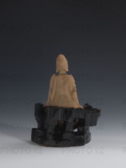 Guanyin Giving Birth to a Son on a Rock; sculptor unknown. Figurine of the goddess made of light coffee-colored alabaster, mounted on a stone base feldspar with red and pink spots, painted with black lacquer. Qing dynasty 18th/19th century, China. Chinese Buddhist subject: Goddess with a rosary in her right hand, holding a child with her left hand. Sculpture; height 18.2 cm, width 13.8 cm., A small carved figure seated on a carved base, viewed from behind, with a smooth light beige material for the figure and a glossy black material for the irregular, blocky base; the figure wears a draped garment with carved decorative patterns and a hood or headdress, and the base has an angular, stacked appearance with chipped edges and a reflective surface; the background is a smooth gradient from dark gray at the top to lighter gray at the bottom, and the visible colors are beige, black, gray, and a few subtle brownish tones on the base.
