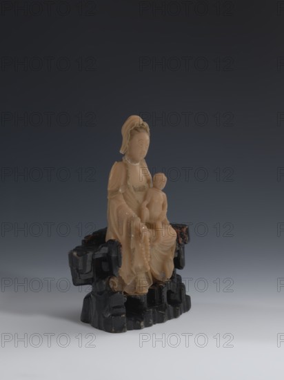 Guanyin Giving Birth to a Son on a Rock; sculptor unknown. Figurine of the goddess Guanyin holding a child with her left hand and a rosary in her right. Made of light coffee-colored alabaster; stone base of feldspar with red and pink spots, painted with black lacquer. Chinese, Qing dynasty 1644–1911, 18th–19th century. Sculpture. Height 18.2 cm, width 13.8 cm. Chinese Buddhism; Chinese art; figurine., A carved beige figure of a seated woman holding a child sits on a carved dark base; the woman wears flowing robes and an elaborate headdress, the child is seated on her lap, and both figures have smooth, rounded facial features and detailed drapery. The base has an irregular, rock-like form with glossy black and dark brown tones. The background is a gradient of dark gray to lighter gray near the bottom, and the surface beneath the object is a pale gray. Colors visible include beige, black, dark brown, dark gray, light gray, and pale gray.