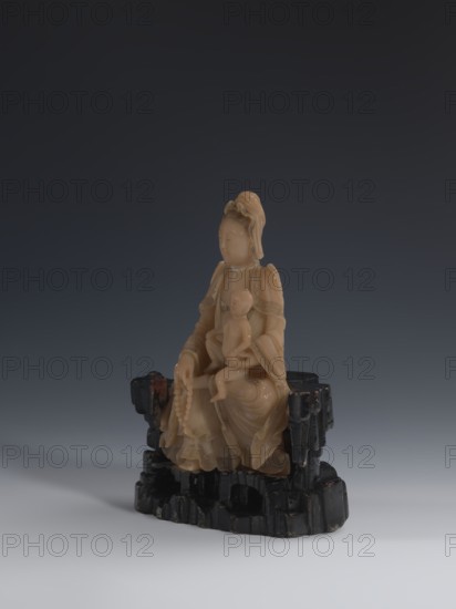 Guanyin Giving Birth to a Son on a Rock; sculptor unknown. Figurine of the goddess made of light coffee-colored alabaster, on a stone base feldspar with red and pink spots, painted with black lacquer. Goddess holds a rosary in her right hand and a child in her left. Chinese; Qing dynasty 1644–1911; 18th–19th century. Sculpture; height 18.2 cm, width 13.8 cm., A carved figure in a warm beige color sits on a dark, glossy black base that resembles a rocky form; the figure appears draped in flowing robes with layered folds and holds a smaller figure on its lap, with a string of beads hanging from one hand and a headpiece with soft curls atop the larger figure's head; the background is a smooth gradient from deep charcoal at the top to pale gray near the bottom.