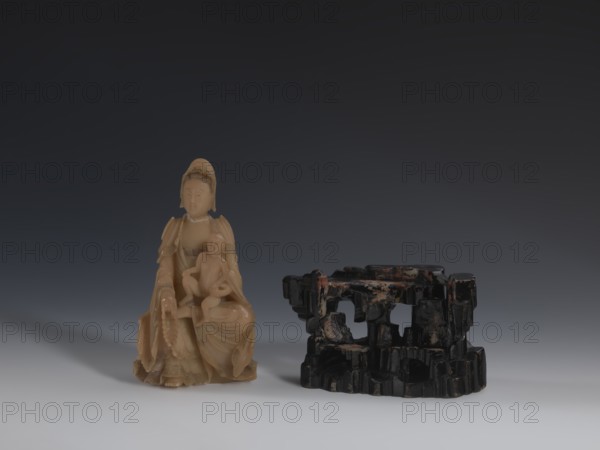 Guanyin Giving Birth to a Son on a Rock, sculptor unknown. Figurine of the goddess made of light coffee-colored alabaster, on a stone base field spar with red and pink spots, painted with black lacquer. Goddess holds a rosary in her right hand and a child in her left. Chinese, Qing dynasty 1644–1911, 18th/19th century. Sculpture; height 18.2 cm, width 13.8 cm., A carved figure in a warm beige color sits on the left, showing a seated person holding a smaller seated figure with flowing drapery and rounded facial features, with the material appearing smooth and slightly translucent; to the right is a dark, irregularly shaped object in black with patches of reddish and brown discoloration, featuring multiple openings and a textured, carved surface; the background transitions from deep charcoal at the top to a lighter gray toward the bottom, and the surface beneath the objects is a pale gray.