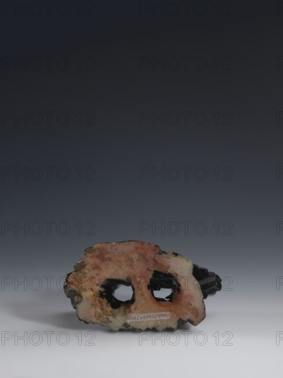 Guanyin giving birth to a son on a rock; sculptor unknown. Figurine of the goddess made of light coffee-colored alabaster, on a stone base feldspar with red and pink spots, painted with black lacquer. Qing dynasty 1644–1911, 18th–19th century, China. Chinese Buddhism; Chinese art. Goddess holds a rosary in her right hand and a child in her left. Sculpture, height 18.2 cm, width 13.8 cm., A roughly horizontal irregularly shaped object sits near the bottom of the image against a gradient background, showing a mix of pink, peach, beige, brown, black, and cream colors on its surface; the object has two rounded openings near its middle and a darker black form visible through and behind those openings, with mottled patches and a slightly translucent appearance in places, and a soft diffuse shadow beneath it on a light gray surface.