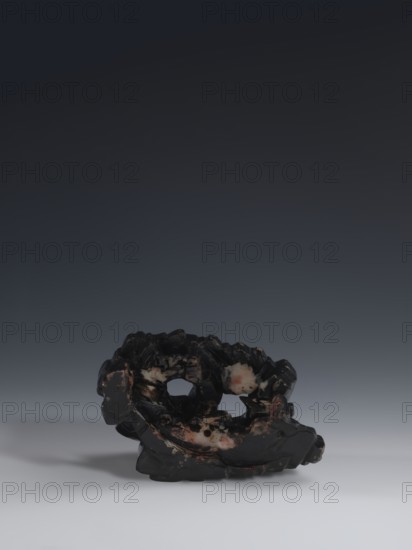 Guanyin Giving Birth to a Son on a Rock; unknown creator; Chinese art; Chinese culture; Qing 1644–1911; Chinese Buddhism; figurine of the goddess with a rosary in her right hand, holding a child with her left hand; made of light coffee-colored alabaster; stone base of feldspar with red and pink spots, painted with black lacquer; 18th–19th century, China; sculpture; height 18.2 cm, width 13.8 cm., A dark, irregularly shaped object with a rough, carved surface sits near the bottom of the image against a smooth gradient background that transitions from very dark at the top to lighter gray toward the bottom. The object is predominantly deep black with patches of pale pink and off-white scattered across its surface, and areas of muted brown and reddish tones. It has multiple openings and hollows, an uneven jagged top, and a rounded base that casts a soft gray shadow on the lighter foreground.