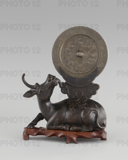 Mirror with TLV decoration on a pedestal in the form of a qilin on a base. Chinese; Qing dynasty 1644–1911. Mirror: cast from chiseled bronze, decoration in flat relief with a centrally placed knob enclosed in a square, four T-shaped motifs attached, and L- and V-shaped motifs at the edge; likely 18th century mirror itself cast from an earlier product, possibly 6th century–?. Unicorn qilin: cast from chiseled bronze. Base: carved wood. Sculpture/mirror; height 26 cm, diameter 16.2 cm., A sculpted reclining animal figure with curved horns and stylized ears, rendered in a dark brown material with lighter brown highlights, rests on a carved wooden base in a reddish-brown color; behind and partly above the figure is a round, ornate disc in a metallic gray-green tone with raised concentric decorative patterns, held by a dark brown decorative support connected to the animal.