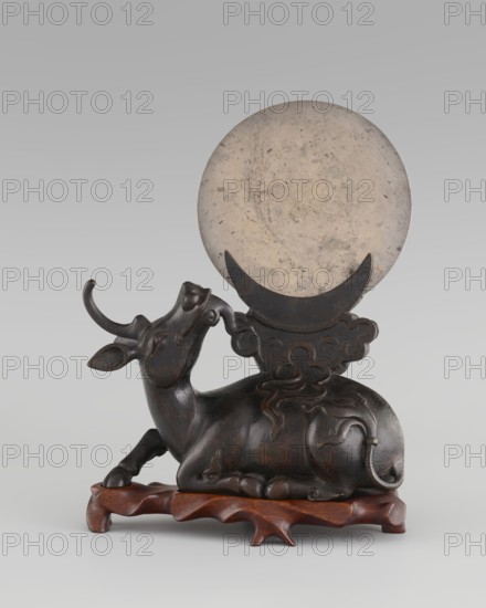 Mirror with TLV decoration on a pedestal in the form of a qilin on a base; Chinese, Qing dynasty 1644–1911. Mirror: cast from chiseled bronze, decoration in flat relief with a centrally placed knob enclosed in a square, four T-shaped motifs attached, and L- and V-shaped motifs at the edge. Pedestal: qilin cast from chiseled bronze; base: carved wood. 18th century mirror cast from an earlier product, possibly 6th century–?. Sculpture/mirror. Height 26 cm; diameter 16.2 cm., A sculpted reclining animal with curved horns rests on a carved wooden base, the animal appearing dark brown to black with a smooth, slightly polished surface and carved decorative lines on its body and tail; behind and above it is a large round disc that is mottled light beige to gray, held by a crescent-shaped support that is darker in color, and the background is a soft gradient of light gray.