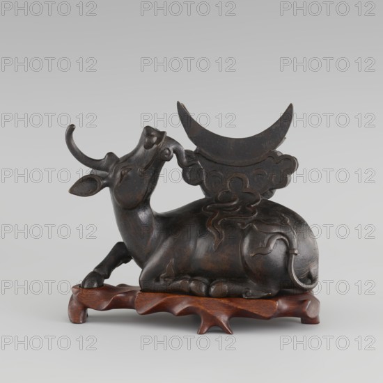 Mirror with TLV decoration on a pedestal in the form of a qilin on a base. Chinese, Qing dynasty 18th century. Mirror: cast from chiseled bronze, decoration in flat relief—centrally placed knob enclosed in a square with four T-shaped motifs attached; L- and V-shaped motifs at the edge. Unicorn qilin: cast from chiseled bronze. Base: carved wood. Mirror possibly cast from an earlier product 6th century - ?. Sculpture/mirror. Height 26 cm; diameter 16.2 cm., A dark brown sculpted figure of a reclining animal with curved horns rests on a carved wooden base; the animal has a smooth, polished surface with subtle lighter brown highlights and carved ornamental lines on its body, and a decorative crescent-shaped element with cloud-like motifs rises from its back in a similar dark brown tone, while the base is a warmer reddish-brown wood.