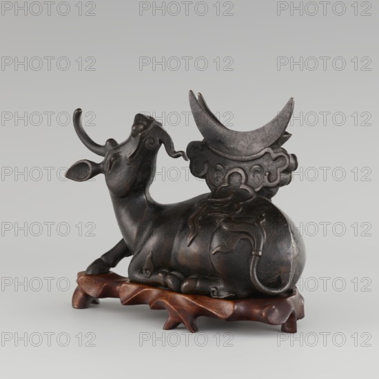 Mirror with TLV decoration on a pedestal in the form of a qilin on a base; Chinese, Qing dynasty 1644–1911; mirror cast from chiseled bronze with decoration in flat relief: centrally placed knob enclosed in a square with four T-shaped motifs attached, and L- and V-shaped motifs at the edge; unicorn qilin cast from chiseled bronze; carved wooden base; 18th century mirror cast from an earlier product, possibly 6th century–?; sculpture, mirror; height 26 cm, diameter 16.2 cm; bronzes, mirrors, castings, qilin iconography., A dark brown sculptural figure of a reclining bovine with curved horns and a raised head sits on a carved reddish-brown wooden base; the bovine has smooth rounded forms, stylized flowing decorative lines along its body and tail, and a large crescent-shaped element rising behind it that rests on a cloud-like carved support, all rendered in shades of dark brown and black with subtle highlights.