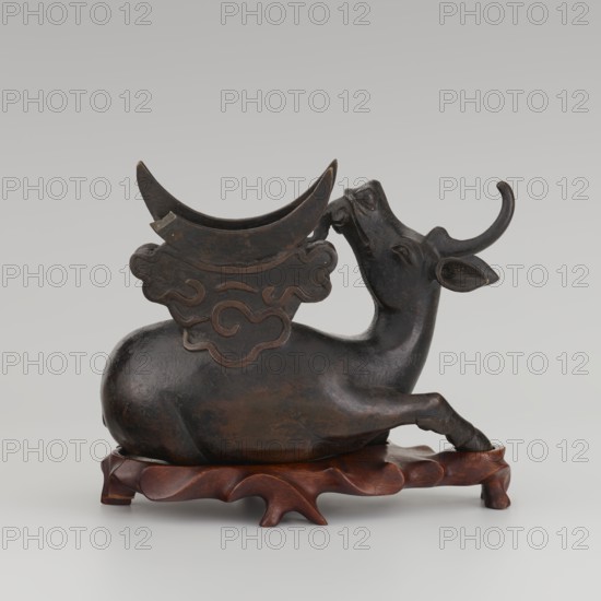 Mirror with TLV decoration on a pedestal in the form of a qilin on a base. Chinese; Qing dynasty 1644–1911. Mirror: cast from chiseled bronze, decoration in flat relief—centrally placed knob enclosed in a square with four T-like motifs attached; L- and V-shaped motifs at the edge. Unicorn qilin: cast from chiseled bronze. Base: carved wood. 18th century mirror cast from an earlier product, possibly 6th century–?. Sculpture, mirror. Height 26 cm; diameter 16.2 cm., A sculpted dark brown animal reclining on a carved reddish-brown wooden base, with a curved horn, elongated neck, and a raised front leg; a decorative crescent-shaped element rests on the animal's back above an ornamental cloud-like plate with carved swirling shapes; the surface shows areas of lighter brown highlights and subtle wear against a neutral gray background.