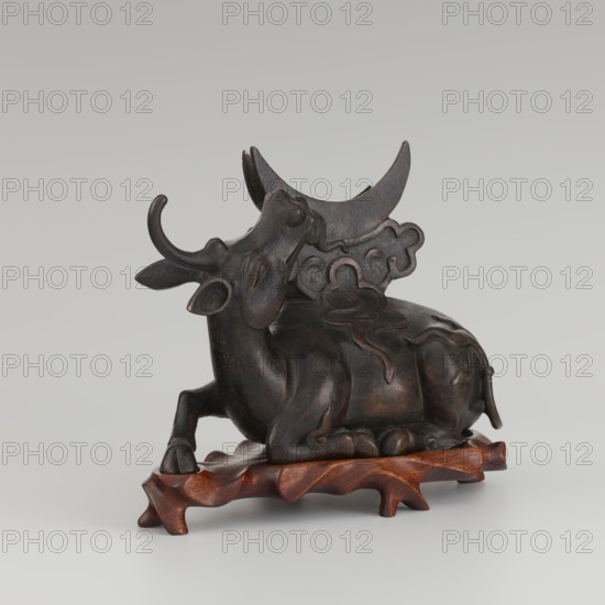 Mirror with TLV decoration on a pedestal in the form of a qilin on a base. Chinese; Qing dynasty 1644–1911. Mirror: cast from chiseled bronze, decoration in flat relief—centrally placed knob enclosed in a square with four T-shaped motifs attached; L- and V-shaped motifs at the edge. Unicorn qilin: cast from chiseled bronze. Base: carved wood. 18th century mirror cast from an earlier product, possibly 6th century–?. Sculpture, mirror. Height 26 cm; diameter 16.2 cm., A dark brown sculpted figure of a reclining bovine with stylized curved horns and carved decorative swirls on its back rests on a polished reddish-brown wooden base; the bovine has a smooth, slightly glossy surface with subtle lighter brown highlights and rounded limbs, and behind its head a crescent-shaped element rises with cloud-like carved motifs, all shown against a pale gray background.