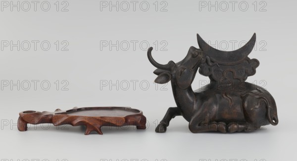 Mirror with TLV decoration on a pedestal in the form of a qilin on a base. Chinese, Qing dynasty, 18th century mirror cast from an earlier product, possibly 6th century onward. Materials: mirror—cast from chiseled bronze with flat-relief TLV decoration: centrally placed knob enclosed in a square with four T-like motifs attached, L- and V-shaped motifs at the edge; unicorn qilin—cast from chiseled bronze; base—carved wood. Sculpture/mirror, height 26 cm, diameter 16.2 cm., A sculpted dark brown object in the shape of a reclining animal with curved horns and decorative raised patterns sits to the right, showing smooth rounded forms and a slightly glossy surface; to the left a low, organic-shaped wooden stand in warm reddish-brown with carved, irregular edges and short legs rests on the same surface. The background is a gradient of pale gray to off-white. Colors visible include dark brown, reddish-brown, gray, black, and off-white.