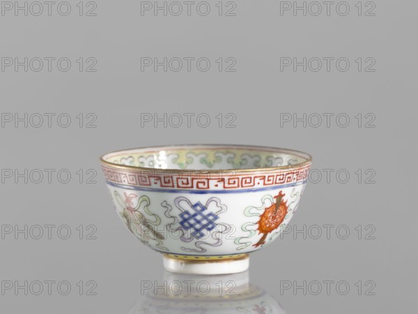 Bowl, gift, Qing dynasty 1644–1911, 19th century, China. Colored porcelain bowl on a foot, height 3.6 cm, diameter 6.6 cm. Decorated on the outside with a meander stripe and Buddhist symbols—including cranes, endless knot—and a lambrequin pattern. Function: vessel; style: Chinese ceramics., A small porcelain bowl with a rounded shape and a short foot, shown against a plain gray background. The exterior of the bowl is white with decorative motifs in several colors including red, blue, green, orange, purple, and gold. A red geometric band runs near the rim, and a thin gold line edges the top. Below the band are stylized cloud-like green and purple swirls and a few emblem-like motifs in blue and orange, with delicate pale green ribbon-like accents. The interior rim shows a repeating pale green scroll pattern with gold edging. The bowl casts a soft reflection on the surface beneath it.