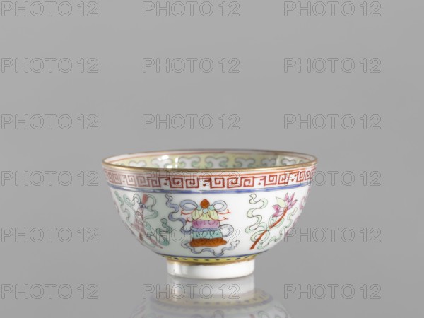 Bowl, Qing dynasty 1644–1911, 19th century, China. Colored porcelain bowl on a foot, height 3.6 cm, diameter 6.6 cm. Decorated on the outside with a meander stripe and Buddhist symbols including cranes and the endless knot, lambrequin pattern. Function: vessel., A small porcelain bowl sits on a reflective surface against a plain gray background, showing a rounded exterior with a narrow base and flared rim. The bowl is primarily white with decorative painted motifs in colors including red, green, blue, pink, orange, yellow, and gold. Around the rim is a band with a red geometric pattern and a thin gold edge. The exterior decoration includes flowing ribbon-like cloud forms in pale green and blue, floral elements in pink and green, and a stack of rounded ornamental shapes in orange, pink, blue, and green tied with a bow. A narrow yellow band with small blue details encircles the foot, and the bowl’s interior rim also shows repeating pale green cloud-like motifs with gold edging. The bowl’...