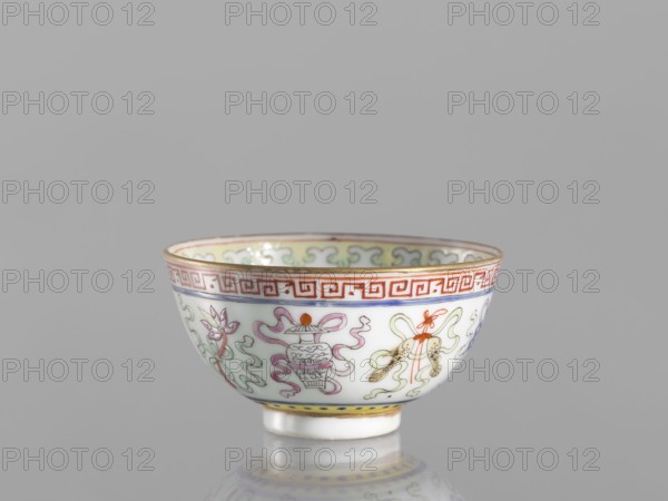 Bowl, gift, Qing dynasty 1644–1911, 19th century, China. Colored porcelain bowl on a foot, height 3.6 cm, diameter 6.6 cm. Decorated on the outside with a meander stripe and Buddhist symbols cranes, endless knot, lambrequin pattern; Chinese ceramics, bowls; related to Boxer Rebellion 1899–1901., A small porcelain bowl with a glossy white surface sits against a plain gray background, decorated around its exterior with a repeating band of geometric motifs in red near the rim, a thin gold trim at the top edge, and a narrow yellow band with blue dots near the foot; the bowl shows painted ornamental designs in soft green, pale pink, lavender, orange, and blue including stylized floral and ribbon-like shapes and a small rendered vessel, with delicate gold accents and a reflective sheen on its curved surface.