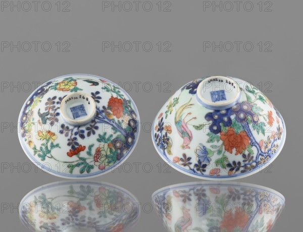 Bowl with a lid, Chinese; Qing dynasty, Qianlong period 1736–1795; colored porcelain; decoration: in a garden setting boulders, trees, peonies, magnolias, chrysanthemums two pheasants and a butterfly; signed; vessel; height 7.5 cm, diameter 11.7 cm, weight 0.2 kg., Two small porcelain bowls are shown upside down on a smooth reflective surface, each decorated with floral and bird motifs. The bowls are primarily white with intricate painted designs in a palette that includes blue, green, red, orange, pink, yellow, and gold accents. One bowl features a prominent red-orange flower and leafy green stems with blue ornamental detailing, while the other shows a colorful bird with pink and green plumage among similar floral patterns and blue branch-like decorations. Both bowls have a ringed foot and a central recessed base area, and their painted surfaces and the gray background are reflected beneath them.