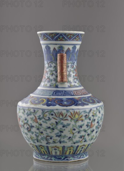 Vase with threads in the shape of bamboo twigs; maker, Chinese, Qing dynasty, Qianlong period 1736–1795; colored porcelain; decoration: lotus tendril on the belly, artemisia leaves, lambrequin ornament on the shoulders, geometric and plant borders on the neck; plant motifs; vessel; height 20 cm, diameter 14.5 cm, weight 0.8 kg., A decorative ceramic vase with a flared neck and bulbous body sits against a gray background, featuring a palette of blue, white, green, yellow, red, and gold; the neck shows stylized leaf motifs in blue and green and a vertical cylindrical red and gold accent attached to its front, while the shoulder and body display scrolling floral vines with lotus-like flowers in red, yellow, and green on a white ground, bordered by geometric and cloud-like bands in blue and red near the top and a band of vertical panel motifs in blue, yellow, and red near the base, with a narrow blue patterned rim at the foot.