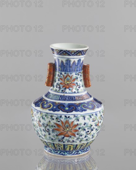 Vase with threads in the shape of bamboo twigs; colored porcelain; Chinese; Qing dynasty, Qianlong period 1736–1795; decoration: lotus tendril and artemisia leaves on the belly; lambrequin ornament on the shoulders; geometric and plant borders on the neck; motifs: bamboos, lotuses, lotus tendril ornament, meander ornament, geometric ornaments; vessel; height 20.0 cm, diameter 14.5 cm; maker, function: vase; subject: flowers., A ceramic vase sits against a plain gray background, featuring a bulbous body, a narrowed neck, and a flared rim, with a pair of short tubular handles on the neck. The surface is decorated with ornate painted floral and foliate motifs in colors including blue, white, orange, green, yellow, and a reddish-brown on the handles, with bands of geometric and curving patterns encircling the neck and shoulder. The primary floral motifs show stylized flowers with orange petals and green leaves amid scrolling blue vines, and the rim and base areas display repeating decorati...