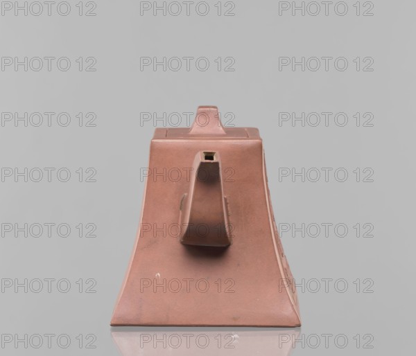 Teapot with a lid, teapot for tea, Harbin China, ceramics, Chinese art, Chinese culture, Qing dynasty 1644–1911, Yixing style, red-brown stoneware, 19th century, China, vessel, height 12.2 cm, width 16.3 cm, inscriptions., A trapezoidal ceramic object in a muted reddish-brown color sits centered on a reflective surface against a pale gray background. The object has a flat top with a small raised rectangular knob, and a central protruding spout-like feature on its front face that is hollow at the tip. The sides slope outward toward the base, and the surface shows subtle variations in tone and a few small blemishes and scuffs in darker and lighter reddish-brown shades. The reflection of the object's base appears faintly on the surface below, and overall the visible colors are reddish-brown and gray with minor darker and lighter reddish-brown marks.