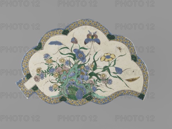 Leaf-shaped saucer, Chinese, Kangxi period 1662–1722; colored porcelain with leaves, butterflies, and plant motifs; vessel; height 1.6 cm, width 26.0 cm, depth 17.0 cm., A scalloped, fan-shaped ceramic plaque sits against a neutral gray background, decorated with a central botanical arrangement of stylized flowers and foliage rendered in shades of blue, green, yellow, pale pink, and cream, with slender green stems and leaves weaving through the composition; a few blue and yellow blossoms, some small daisy-like flowers, and several seedpod shapes are visible, along with a textured rock-like form near the base in green and blue. The plaque’s rim features a repeating geometric pattern in yellow and blue framed by a narrow blue border, with small dark green cloud-like motifs at intervals; the plaque surface itself is a warm off-white or cream with fine speckling.