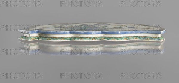 Leaf-shaped saucer, Chinese, Kangxi period 1662–1722; colored porcelain with leaves, butterflies, and plant motifs; vessel; height 1.6 cm, width 26.0 cm, depth 17.0 cm., A low-profile decorative ceramic object viewed in profile resting on a reflective surface, showing an undulating scalloped edge with a glossy blue rim and a white band beneath, a patterned green band with yellow floral motifs along the lower edge, and a pale top surface with muted multicolored painted decoration; the reflection on the surface mirrors the colors and shapes.