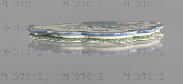Leaf-shaped saucer, Chinese; Kangxi period 1662–1722; colored porcelain; decorated with leaves, butterflies, and plant motifs; vessel; height 1.6 cm, width 26.0 cm, depth 17.0 cm., A low-profile ceramic object with a scalloped edge sits on a reflective surface, showing a thin layered construction with a blue rim on top, a white middle layer, and a patterned green band beneath; the top surface includes floral motifs in muted greens and blues against a pale background, and the decorative band shows repeating leaf-like shapes in green with yellow accents, all reflected softly on the gray surface below.