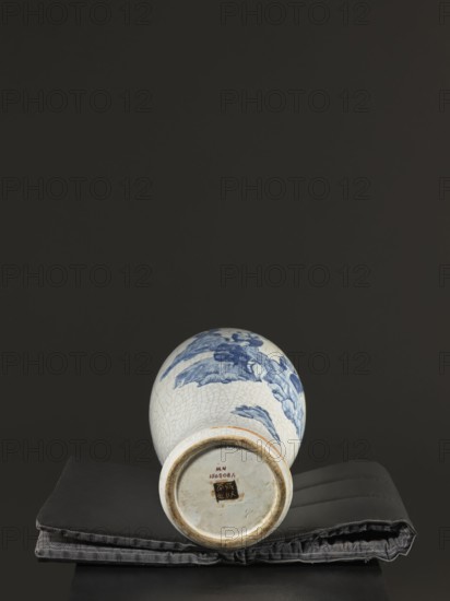 Vase, figurative scenes, ceramics, gardens, Chinese art, Chinese culture, blue-and-white porcelain, games, cracklework decoration, play, Qing 1644–1911, 18th–19th century Jiaqing period, 1797–1820, Qing dynasty, China, vessel, height 54.5 cm, width 9.8 cm, A ceramic vase lying on its side on a folded fabric pad, the vase showing a crackled white glaze with blue painted motifs and a rounded base with an unglazed foot rim, the fabric pad appearing dark gray and black with stitched seams, all set against a solid dark background with shades of black and deep gray.