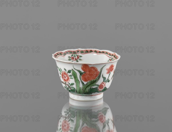 Bowl, porcelain with colorful overglaze enamels in Kangxi style Qing dynasty, 1662–1722. Decorated with floral motifs, plum trees, and bird images. Vessel, height 5.4 cm, diameter 8.8 cm; China., A small white porcelain cup with a scalloped rim sits on a reflective surface against a gray background; the cup is decorated with floral motifs in red and green, including a few large red flowers and several green leaves, and a patterned red-and-green border runs just inside the rim, with the cup’s reflection visible beneath it.