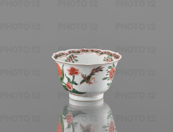 Bowl, vessel, ceramics, colorful decorations, Kangxi style, colorful porcelain with floral pattern, plum trees and bird imagery, Qing dynasty 1644–1911, Kangxi period 1662–1722, decorated with floral motifs, China; height 5.4 cm, diameter 8.8 cm., A small ceramic cup with a scalloped rim sits on a reflective surface, showing its mirrored underside; the cup is primarily white with painted floral and bird motifs in red and green, with touches of pale orange and dark green detailing, and a thin red line near the base.