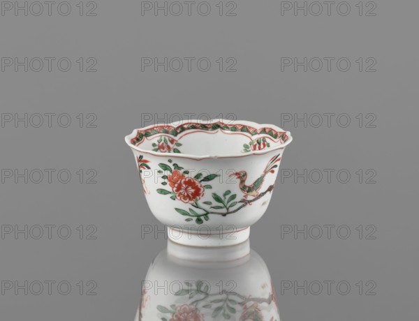 Bowl, ceramic vessel with colorful Kangxi-style porcelain decorations; floral motifs including plum trees and bird images; Qing dynasty Kangxi period, 1662–1722; China; height 5.4 cm, diameter 8.8 cm., A small white porcelain cup with a scalloped rim sits against a gray background, decorated with red and green floral motifs and a green-and-red bird perched on a brown branch, with additional red and green decorative patterns along the inner rim and a pale reflection of the cup visible on the glossy surface below.