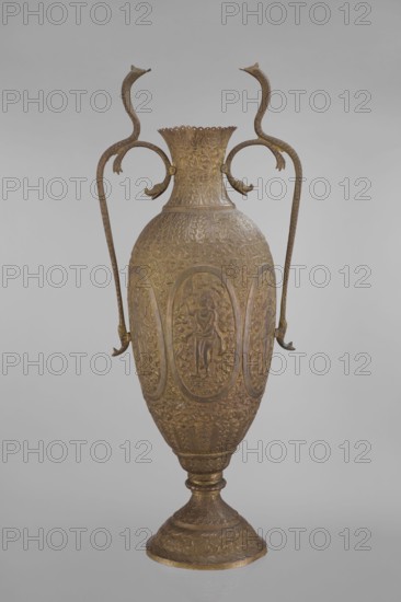 Vase, repossession; Vishnu; Indian art; Indian culture; cobras snakes; plant ornament; geometric ornament; figural scenes; fish; halos; maces; religion; snakes; processions; flutes; clubs. On the belly six ovoid reserves with figural decoration four female figures with wreaths, two images of Vishnu; plant and animal motifs on the entire surface. 19th century, India. Vessel. Height 65.0 cm; width 27.5 cm; weight 2.95 kg., The image shows an ornate metal vase with a tall, elongated body and a flared neck, finished in shades of gold and bronze; the surface is covered in detailed raised decoration including repeated oval panels with human figures and elaborate floral and foliate patterns; a pair of long, curved handles rise from the shoulders and loop upward with stylized animal head terminals, matching the gold-bronze coloration; the vase stands on a pedestal base that echoes the decorative motifs and colors; the background is a smooth, neutral gray.