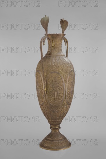 Vase, repossession Kraków - Vishnu, cobras snakes, plant ornament, geometric ornament, figural scenes, fish, halos, maces, religion, snakes, processions, flutes, clubs. On the belly six ovoid reserves with figural decoration four female figures with wreaths, two images of Vishnu; plant and animal motifs on the entire surface. 19th century, India, vessel; height 65.0 cm, width 27.5 cm, weight 2.95 kg., A tall ornate vase with a metallic golden-brown surface stands against a plain light-gray background; the vase has an elongated ovoid body on a flared pedestal base and features intricate raised decorative patterns covering its surface, including repeated oval panels with sculpted human figures and surrounding floral and foliate motifs, and a narrow neck with scalloped rim; two long curved handles rise from the shoulder to the neck and end in stylized serpent or cobra heads, all sharing the same golden-brown hue with subtle darker and lighter variations.