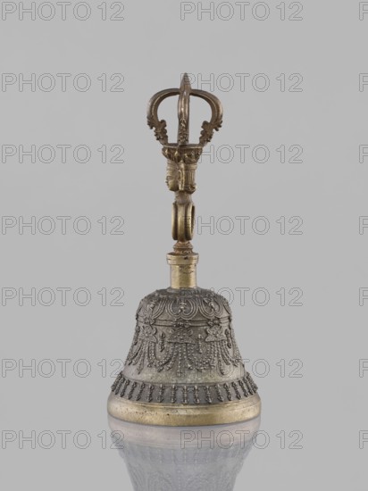 Bell ghanta, purchased; art of Tibetan Buddhism; used in religious rites; ritual object bell/vajra; features heads, mantras; percussion idiophone; bowl decorated with festoons; 19th century; Tibet; height 18.7 cm, width 8.5 cm, A metal bell with a decorative handle sits against a plain light gray background, the bell and handle reflected faintly on a glossy surface below; the bell body is dark gray with ornate raised patterns and textural detailing and a band of aged golden color at its base, while the handle above is golden-bronze with sculpted elements including a small humanlike head and an open crownlike top, showing tones of gold, bronze, and dark gray.