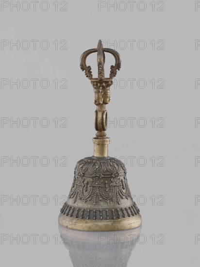 Bell ghanta, 19th century, Tibet. Ritual object used in Tibetan Buddhist rites; percussion idiophone. Metal bell with decoration of the bowl in the form of festoons. Includes vajra head and mantras. Height 18.7 cm, width 8.5 cm., A decorative metal bell with a dark metallic body and golden accents sits upright against a plain light gray background, showing intricate raised ornamental patterns on the bell's surface and a rounded base with a gold-toned rim; its handle is an ornate vertical column topped by a sculpted crown-like element and a small carved head, all in a darker gold to bronze hue, and the bell's lower portion is reflected faintly on the surface beneath, with visible colors including gray, dark metallic, gold, and bronze.