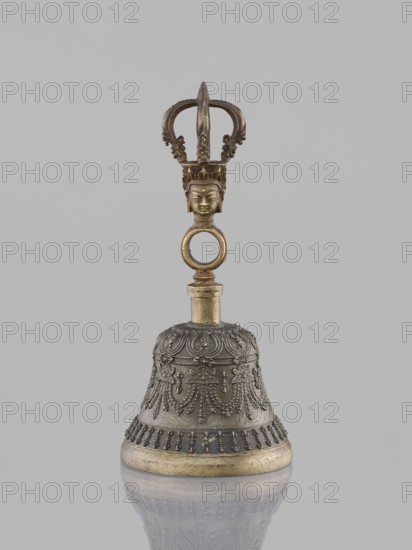 Bell ghanta, 19th century, Tibet. Ritual object used in Tibetan Buddhist rites; percussion idiophone bell with vajra/heads and mantras. Decoration: bowl decorated with festoons. Height 18.7 cm; width 8.5 cm., The image shows a decorative metal bell with a patinated finish in shades of gold and dark gray; the bell rests on a reflective surface and features ornate raised patterns on its dark gray body and a gold-colored rim, with a gold-colored cylindrical neck leading to a handle topped by a sculpted head and a loop and forked crown-like element, all against a plain light gray background.