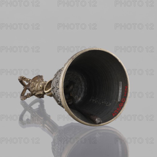 Bell ghanta, 19th century, Tibet. Ritual object used in Tibetan Buddhist religious rites; percussion idiophone. Materials and features: bell with decoration of the bowl in the form of festoons; includes vajra dorje head, mantras. Height 18.7 cm; width 8.5 cm., A metal bell lying on its side against a gray background, showing a dark interior and a rim with a metallic edge; the exterior near the rim has ornate raised decoration and a textured band, and a decorative handle extends from the bell with an intricate openwork design; a clapper is visible inside the bell, and the surface beneath the bell produces a reflective mirror-like reflection; visible colors include gray, dark gray, black, bronze, and hints of red.