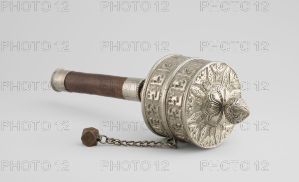 Prayer wheel, Tibetan Buddhist art, Himalayan culture, Himalayan art, scrolls, Buddhist art, 19th century, Tibet, cult object, height 26.5 cm, An ornate handheld object lies on a neutral background, showing a cylindrical carved metal head with raised floral and geometric motifs and a rounded, petal-like protrusion; the head is silvery in color with darker shadowed recesses. Attached to the head is a short metal chain leading to a small faceted weight that is brownish in color. Extending from the head is a wooden handle that is brown with visible grain and a slightly darker patina, capped at the end with a silvery metal band. The overall scene contains colors of silver, brown, and subtle gray tones in the background.
