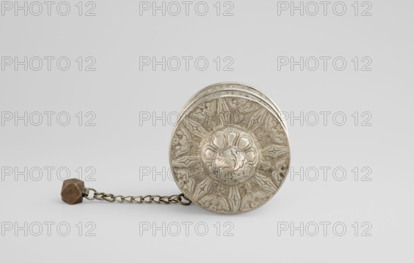 Prayer wheel, Tibetan Buddhist art, Himalayan, scrolls, Buddhist art, 19th century, Tibet, cult object, height 26.5 cm, A round metallic object with ornate raised floral and geometric decoration sits on a pale surface, connected by a short metal chain to a small faceted pendant; the object and chain are mostly silver and bronze in color, the pendant showing brown tones, and the background and surface are shades of light gray.