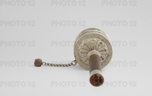 Prayer wheel, Tibetan Buddhist art, Himalayan, Buddhist art, 19th century, Tibet, scrolls, cult object, height 26.5 cm, An object with a cylindrical wooden handle in brown and metal fittings in silver lies horizontally against a light gray background, the handle attached to an ornate circular metal wheel decorated with engraved floral and geometric patterns in silver, a short metal chain in brass tones extends from the wheel to a small faceted weight in brownish bronze, and there are touches of darker brown and a bit of reddish discoloration visible at the end of the handle.
