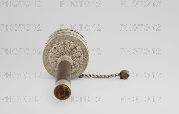 Prayer wheel, Tibetan Buddhist art, Himalayan, Himalayan art, scrolls, Buddhist art, 19th century, Tibet, cult object, height 26.5 cm, An object lying on a light gray surface with a pale gray background, showing a cylindrical wooden handle in brown with metal fittings in silver at one end and a decorative silver circular disk with engraved patterns at the other; attached to the disk is a short metal chain in brass tone ending in a faceted brownish geometric weight; colors visible include brown, silver, brass, pale gray, and a hint of reddish-brown at the far end of the handle.