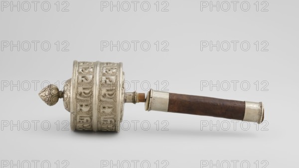 Prayer wheel, repossession, Tibetan Buddhist art, Himalayan culture, Himalayan art, scrolls, Buddhist art, 19th century, Tibet, cult object, height 26.5 cm, A horizontal handheld object with a cylindrical metallic drum on the left and a wooden handle on the right, the drum showing ornate raised decorative patterns and a pointed finial, the metal appearing silver in color and the handle appearing brown with silver-colored metal bands at each end, set against a light gray background.