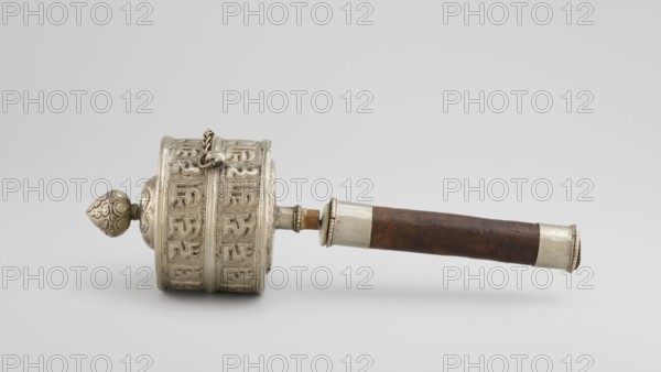 Prayer wheel, Tibetan Buddhist art, Himalayan art, scrolls, Buddhist art, 19th century, Tibet, cult object, height 26.5 cm, A horizontal handheld object with a cylindrical silver metal drum at one end and a brown wooden handle at the other, the metal drum featuring ornate raised decorative patterns and a rounded finial with embossed designs, a short metal chain attached near the top of the drum, silver metal bands and collars connecting the drum to the handle and capping the handle end, and visible colors including silver metallic, brown wood tones, and neutral gray background.