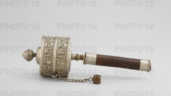 Prayer wheel, Tibetan Buddhist/Himalayan art, 19th century, Tibet. Scrolls, Buddhist cult object, height 26.5 cm., A single handheld cylindrical object rests horizontally on a pale gray background, featuring a metal drum-like end with ornate raised patterns and a pointed finial in metallic silver tones, a dark brown wooden handle with metallic silver bands at each end, and a short metal shaft connecting the drum to the handle; a small chain of linked metal loops with a faceted brown weight hangs beneath the drum. The visible colors include silver, dark brown, brown, and pale gray.