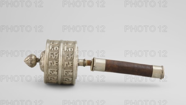 Prayer wheel, Tibetan Buddhist art, Himalayan, Buddhist art, 19th century, Tibet, scrolls, cult object, height 26.5 cm, A handheld cylindrical object rests horizontally on a light neutral background, composed of a decorative metallic drum-like wheel at one end and a wooden handle extending from it; the wheel is silvery with raised patterned motifs and a rounded finial on its outer face that is also silvery and ornamented, while the handle is brown wood with silvery metal bands near both ends and a darker brown section along its length, and there are subtle soft shadows beneath the object.