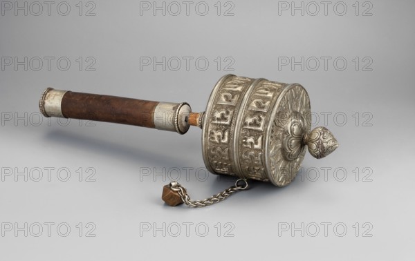 Prayer wheel, Tibetan Buddhist/Himalayan art, Buddhist cult object, scrolls, 19th century, Tibet, height 26.5 cm, A handheld cylindrical object with an ornate metal drum and a wooden handle, resting on a smooth light gray surface against a pale gray background. The drum portion is metallic silver with raised decorative bands and floral-like embossing, and it has a rounded ornamental finial at one end with intricate patterns. The handle is brown wood with silver metal caps at both ends and a short exposed wooden section connecting to the drum. A short silver chain with a small brown faceted weight is attached to the drum and lies on the surface. The visible colors are silver, brown, and shades of gray.