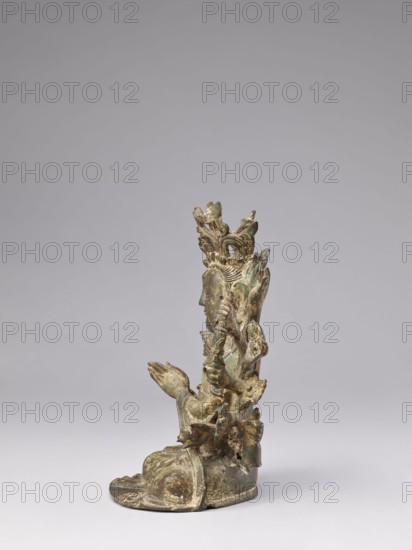 Cundi, unknown sculptor, Chinese/Tibetan, Qing dynasty 1644–1911, 18th–19th century. Sculpture depicting the bodhisattva Cundi with lotus motifs, mudras, sutra, fish, and protective deity/goddess iconography; associated with Buddhism/Chinese Buddhism. Materials not specified. Dimensions: height 24.3 cm, width 22.0 cm, weight 3.38 kg., A small statuette of a seated figure shown in profile against a plain gray background, with a greenish-brown patina and areas of darker and lighter wear, wearing an ornate multi-tiered crown or headdress with floral and leaf-like motifs, a detailed carved face and elongated ear, folded hands held near the chest, layered drapery around the legs and base, and textured decorative elements and small protrusions along the back and shoulders.