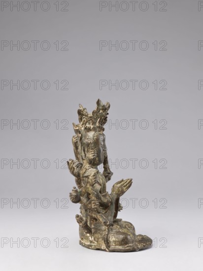 Cundi, unknown sculptor; Chinese/Tibetan Buddhist sculpture, Qing dynasty 18th–19th century; bodhisattva Cundi depicted with lotuses, mudras, sutra, fish; goddess/protective deity iconography; materials not specified; height 24.3 cm, width 22.0 cm, weight 3.38 kg; Chinese culture, Tibetan culture; Buddhism/Chinese Buddhism., A small sculptural figure cast in a dark metallic material with areas of greenish patina and lighter worn highlights, shown in profile and seated on a rounded base; the figure has an elaborate multi-tiered headdress with ornate curving elements, a serene facial expression, and multiple arms arranged around the torso with several hands in different gestures, some held together in a prayer-like pose; the surface shows textured detailing on clothing and adornments, and the overall colors visible are dark bronze, greenish patina, and lighter metallic worn spots against a neutral pale gray background.