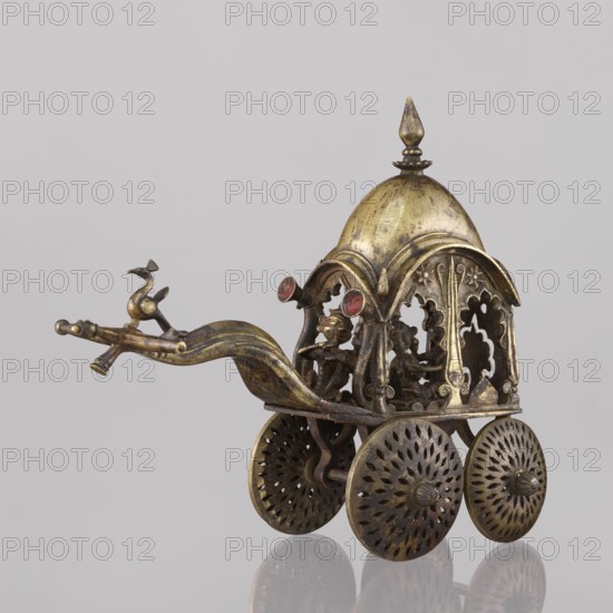 Miniature of a chariot, 19th century, India, model; materials not specified. Depicts a bird perched on a bow-shaped shaft and peacocks; under a canopy topped with a pike sit figures: Krishna feeding on butter; in front, in the driver's seat, there is also Krishna. Height 24.0 cm; width 28.0 cm., The image shows a small ornate metal chariot-like object in gold and bronze tones with a domed canopy topped by a pointed finial, a curved shaft extending forward, and a decorative bird-like figure at the end of the shaft; under the canopy a group of sculpted human-like figures is visible through ornate arched openings, and the vehicle rests on a set of intricately pierced round wheels in matching metallic hues, all set against a plain light gray background with reflections visible beneath the wheels.