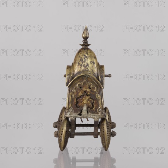 Miniature of a chariot; maker, Indian; 19th century; model; temple chariot; subject: peacocks and birds; a bird sits on a bow-shaped shaft; under a canopy topped with a pike sit figures: Krishna feeding on butter; in front, in the driver's seat, there is also Krishna; height 24.0 cm, width 28.0 cm., A small ornate metal object resembling a wheeled carriage viewed from the front, featuring a domed top with a pointed finial, decorative scalloped and floral cutouts on the front panel, a pair of perforated round wheels on each side connected by an axle, and a mix of metallic tones including gold, brass, brown, and darker tarnished areas against a light gray background.