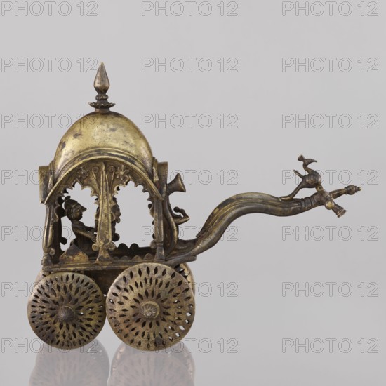 Miniature of a chariot, Indian, 19th century, South Asia; model of a temple chariot with peacocks and birds. A bird sits on a bow-shaped shaft. Under a canopy topped with a pike sit figures: Krishna feeding on butter; in front, in the driver's seat, there is also Krishna. Materials not specified. Height 24.0 cm; width 28.0 cm., A small ornamental wheeled carriage made of metal with a domed roof and a pointed finial, showing metallic tones of gold and bronze; the carriage has arched open sides with decorative scalloped edges and carved column-like supports; inside the carriage a small seated figure is visible in silhouette; the carriage rests on a group of ornate spoked wheels with perforated floral patterns; a curved handle extends from the front with a stylized animal-shaped finial at its end, also in gold and bronze hues; the background is a plain light gray.